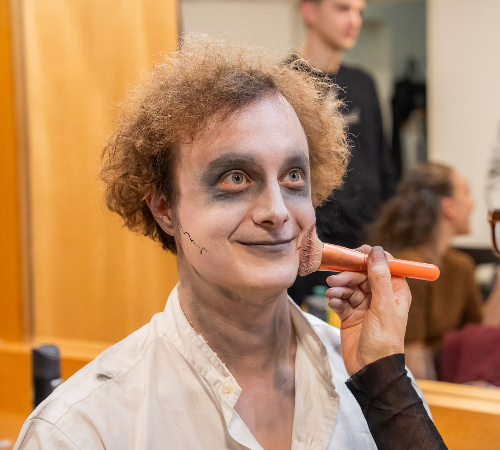 Male student getting makeup done, smiling during an event at The New Academy Musical Theatre Vienna.
