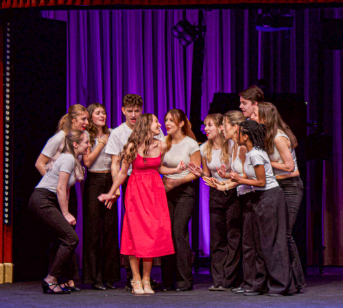 Group of students surrounding a female student in a red dress during a performance at The New Academy Musical Theatre Vienna.